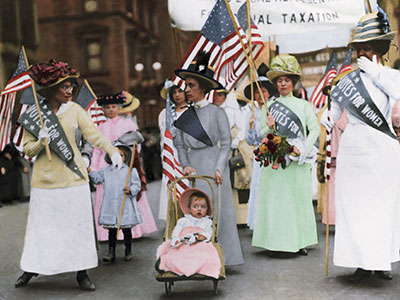 A group of women in early 20th-century clothing march in a suffrage parade. They carry American flags and wear sashes reading &quot;Votes for Women.&quot; One woman pushes a stroller with a child. A banner is visible in the background.
